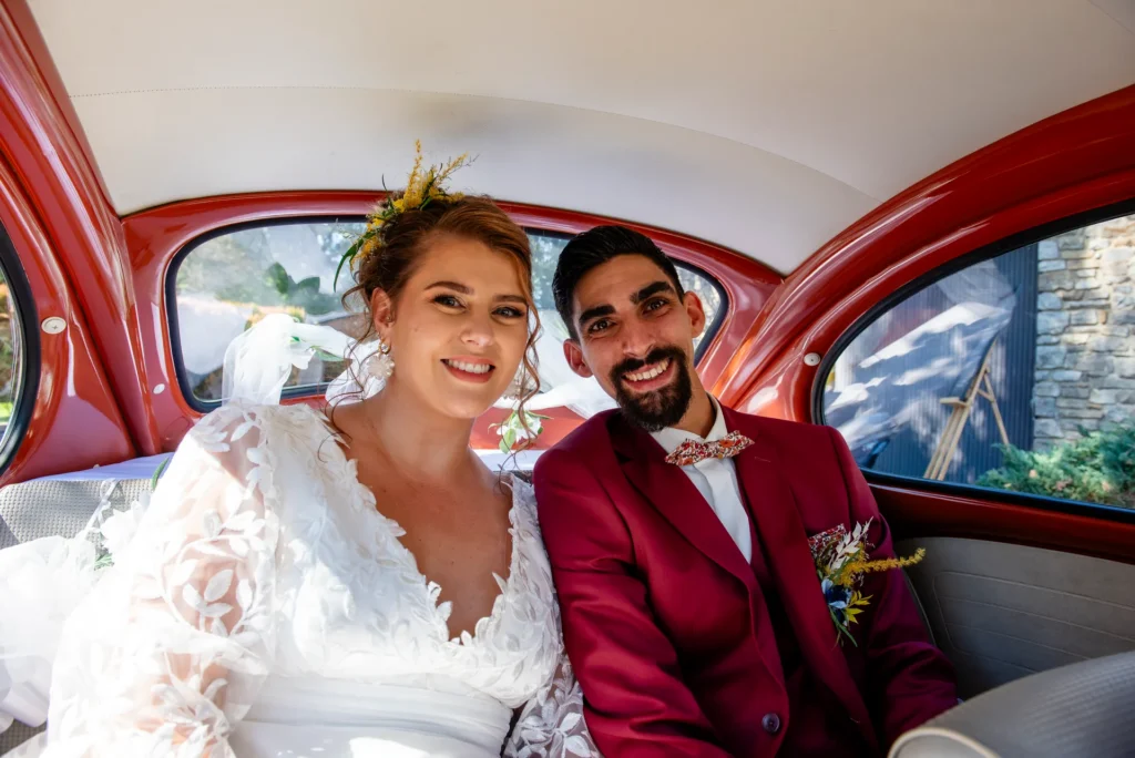 Mariés souriants à l'arrière d'une Coccinelle rouge vintage décorée, arrivée au Domaine de la Raimonderie en Ille-et-Vilaine.